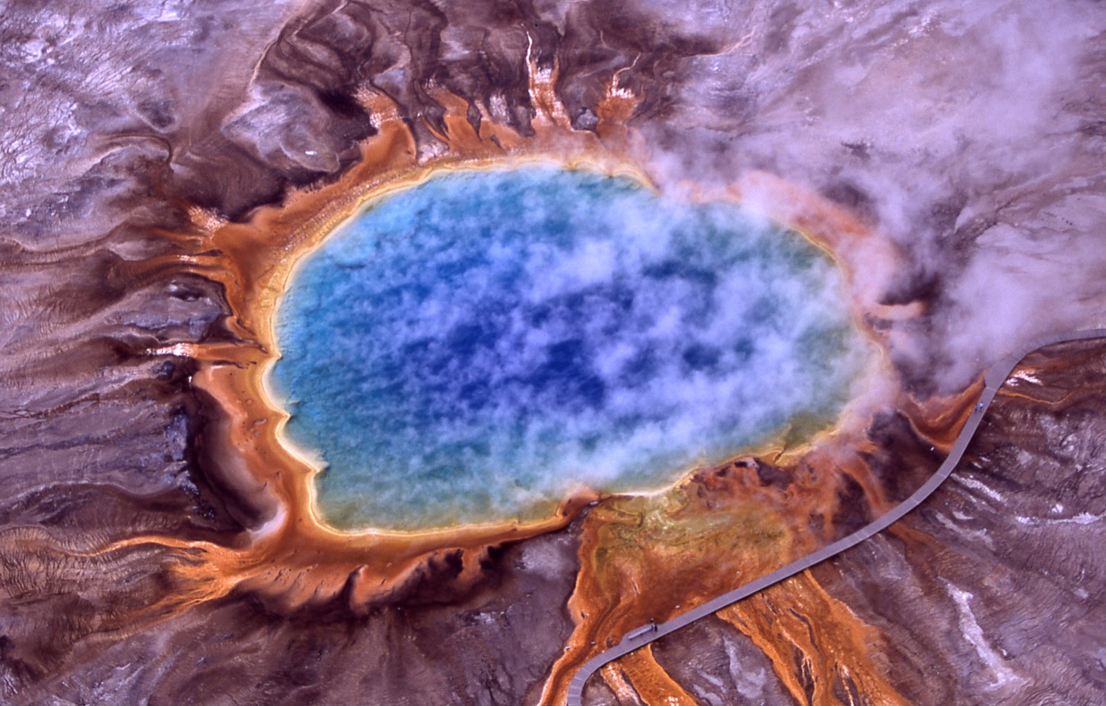 Aerial view of Grand Prismatic Spring; Hot Springs, Midway &amp; Lower Geyser Basin, Yellowstone National Park. The spring is approximately 250 by 300 feet (75 by 91 m) in size. This photo shows steam rising from hot and sterile deep azure blue water (owing to the light absorbing overtone of an OH stretch which is shifted to 698 nm by hydrogen bonding  ) in the center surrounded by huge mats of brilliant orange algae, bacteria and archaea. The color of which is due to the ratio of chlorophyll to carotenoid molecules produced by the organisms. During summertime the chlorophyll content of the organisms is low and thus the mats appear orange, red, or yellow. However during the winter, the mats are usually dark green, because sunlight is more scarce and the microbes produce more chlorophyll to compensate, thereby masking the carotenoid colors. | Yellowstone National Park in United States
